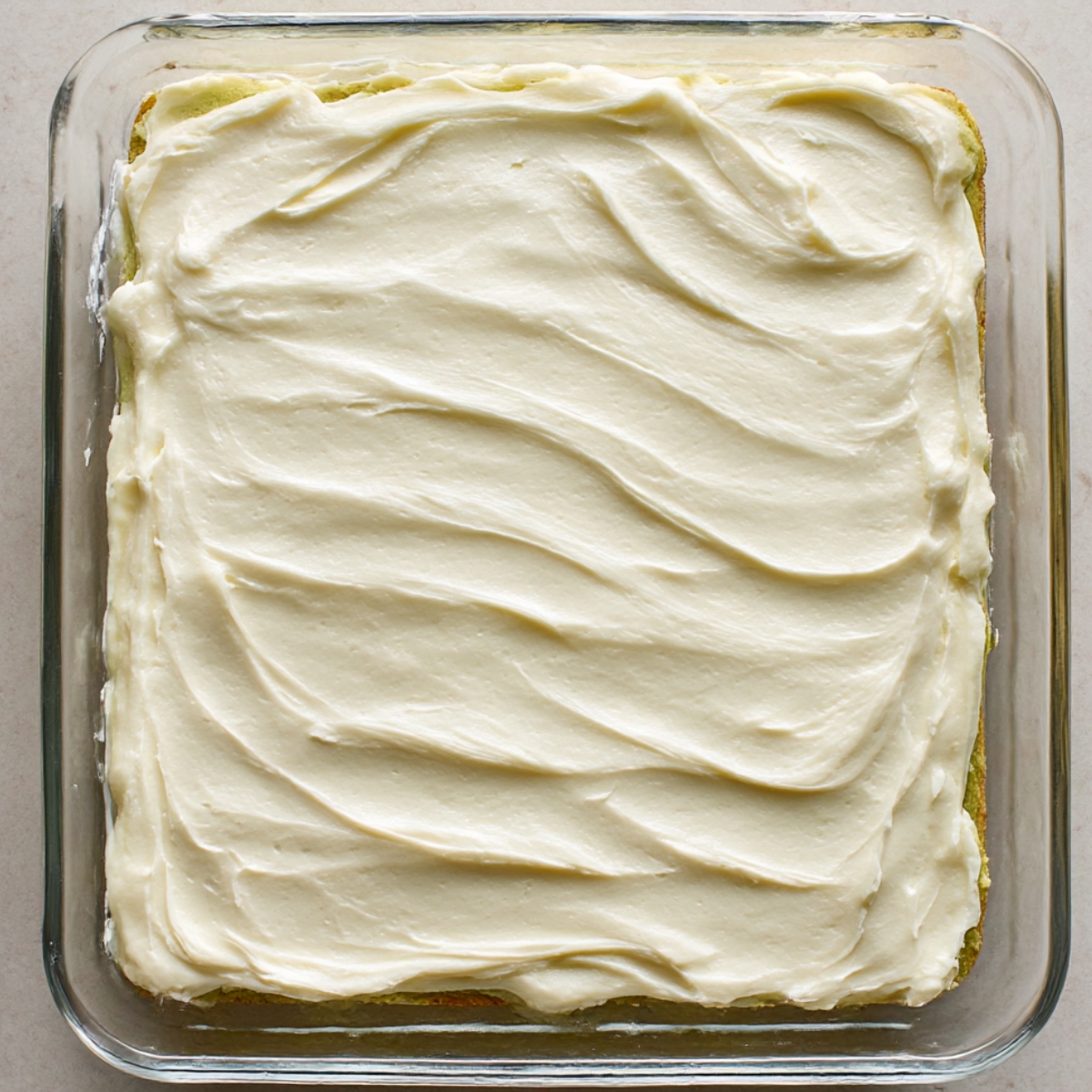 Top view of freshly baked pistachio cookie bars in a rectangular glass dish, covered with a smooth layer of creamy white frosting, ready to cut and serve.