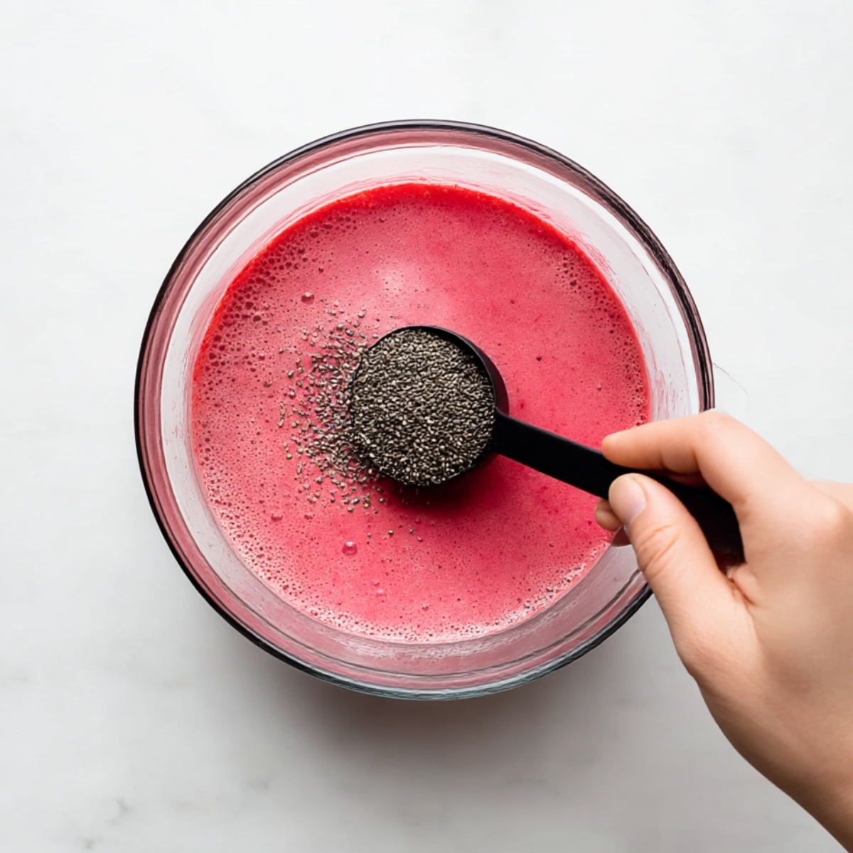 Hand holding a black measuring spoon adding chia seeds into a bowl of vibrant pink raspberry mixture, preparing to make raspberry chia pudding on a white surface.