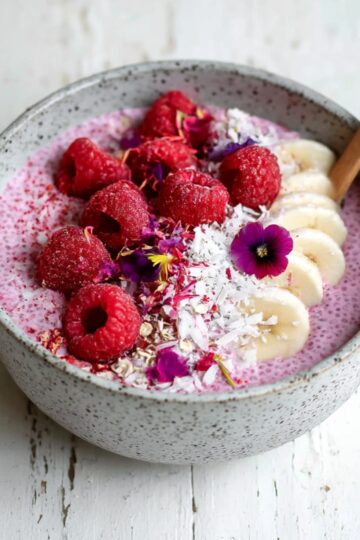 A bowl of vibrant raspberry chia pudding topped with fresh raspberries, banana slices, shredded coconut, oats, and colorful edible flowers, served in a speckled gray bowl with a wooden spoon on a rustic white wooden surface.