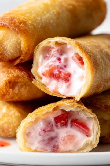 Golden-brown strawberry cheesecake egg rolls filled with creamy cheesecake and fresh strawberry pieces, stacked on a white plate, perfect for a sweet dessert treat.