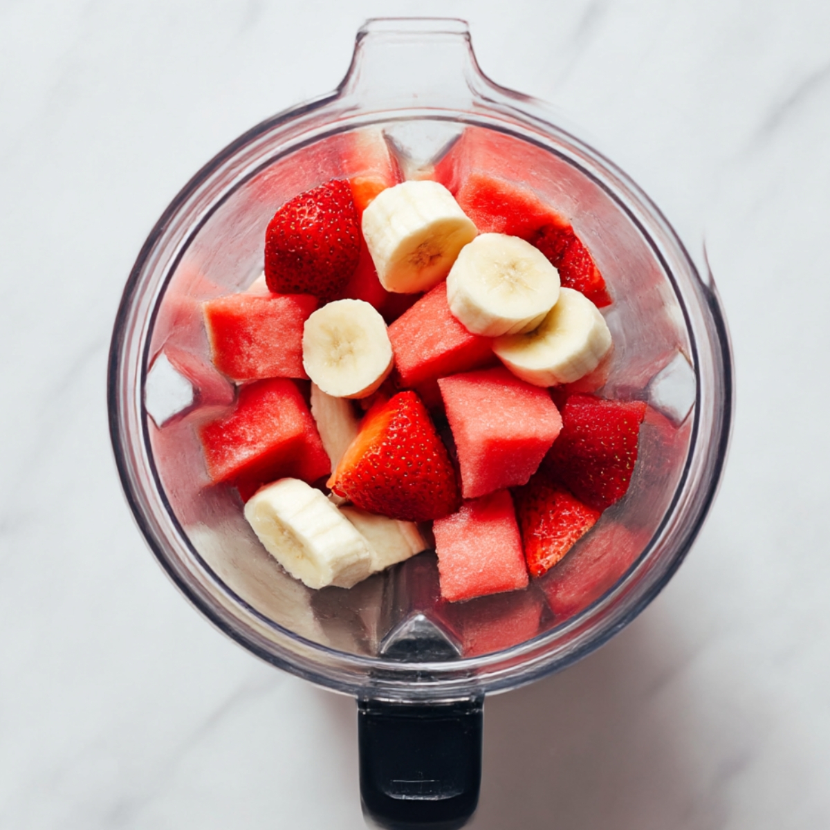 Top view of a blender filled with fresh strawberries, watermelon cubes, and banana slices, perfect for making a refreshing strawberry watermelon smoothie.