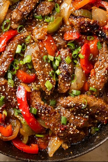 Close-up of Chinese pepper steak stir-fry with tender beef strips, red and green bell peppers, onions, sesame seeds, and green onions in a savory sauce, served in a cast-iron skillet.