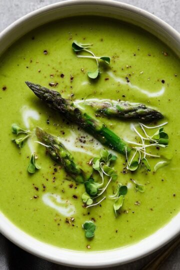 Bowl of creamy asparagus soup garnished with roasted asparagus tips, microgreens, a drizzle of cream, and freshly cracked black pepper, perfect for a healthy and comforting meal.
