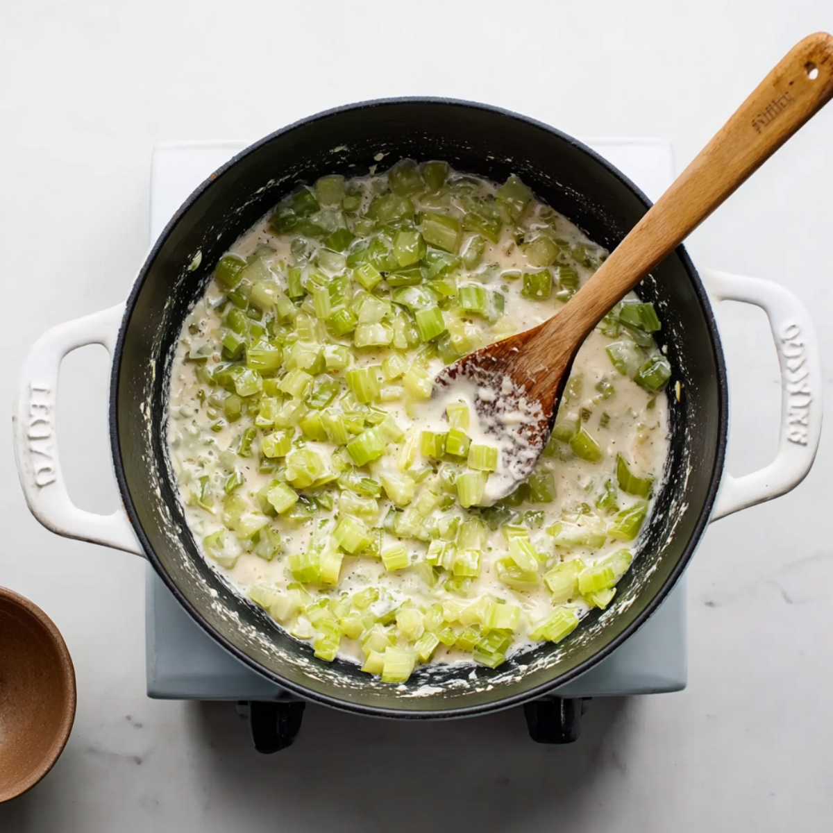 A black pot on a stove with a creamy mixture of chopped celery being stirred with a wooden spoon, showing the preparation stage for a creamy shrimp bisque.