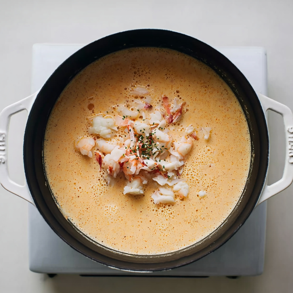 A black pot on a stove filled with creamy shrimp and crab bisque, garnished with chopped seafood and herbs, showcasing a rich, orange-colored seafood soup.