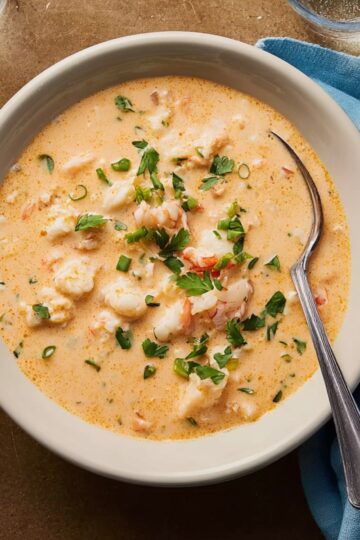 A bowl of creamy shrimp and crab bisque garnished with fresh parsley and green onions, served with a side of crusty bread, showing a rich, orange-colored seafood soup.