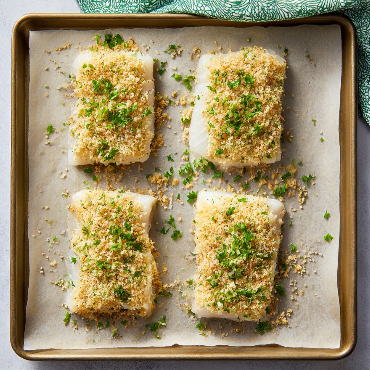 Overhead view of four uncooked cod fillets on a parchment-lined baking sheet, topped with a golden Parmesan breadcrumb crust and sprinkled with fresh parsley, ready for baking.