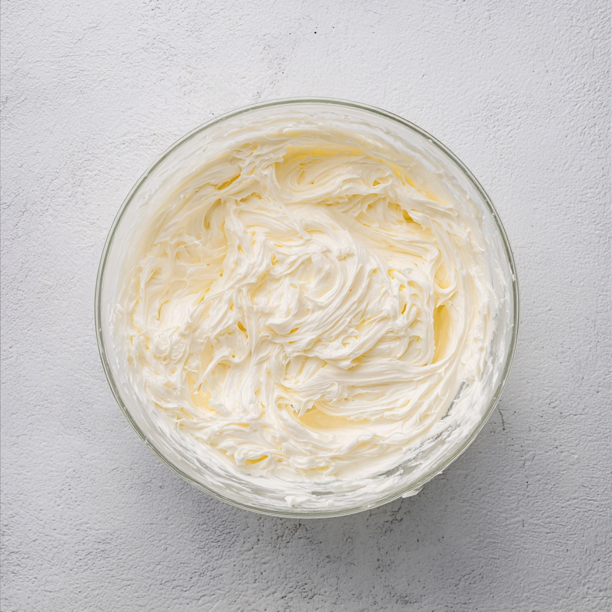 A glass bowl filled with creamy, whipped cream cheese frosting, ready to spread on banana bread brownies, sitting on a light textured surface.