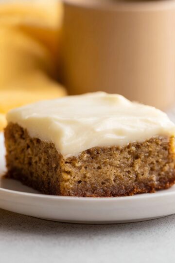 A square slice of moist banana bread brownie topped with creamy white cream cheese frosting, served on a white plate with a soft yellow background.
