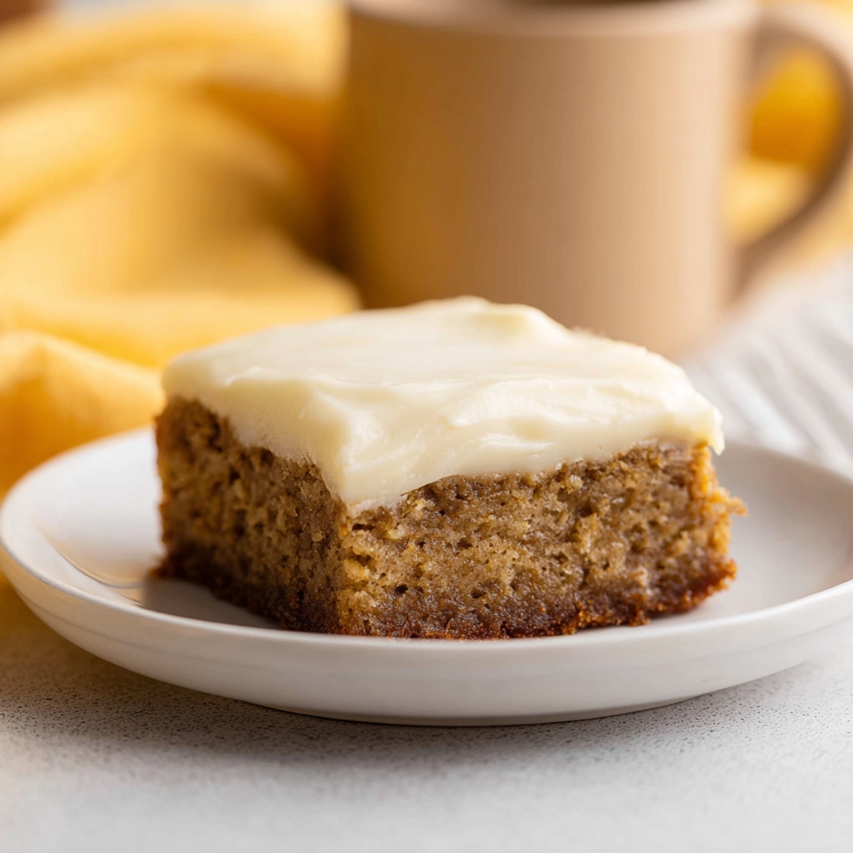 A square slice of moist banana bread brownie topped with creamy white cream cheese frosting, served on a white plate with a soft yellow background.