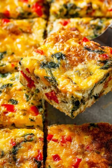 Golden baked make ahead breakfast casserole cut into squares, loaded with melted cheese, fresh spinach, and diced red peppers, perfect for a quick and hearty morning meal.