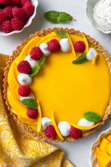 Top view of a bright yellow mango curd tart in a coconut crust, decorated with fresh raspberries, mango slices, dollops of whipped cream, and mint leaves.
