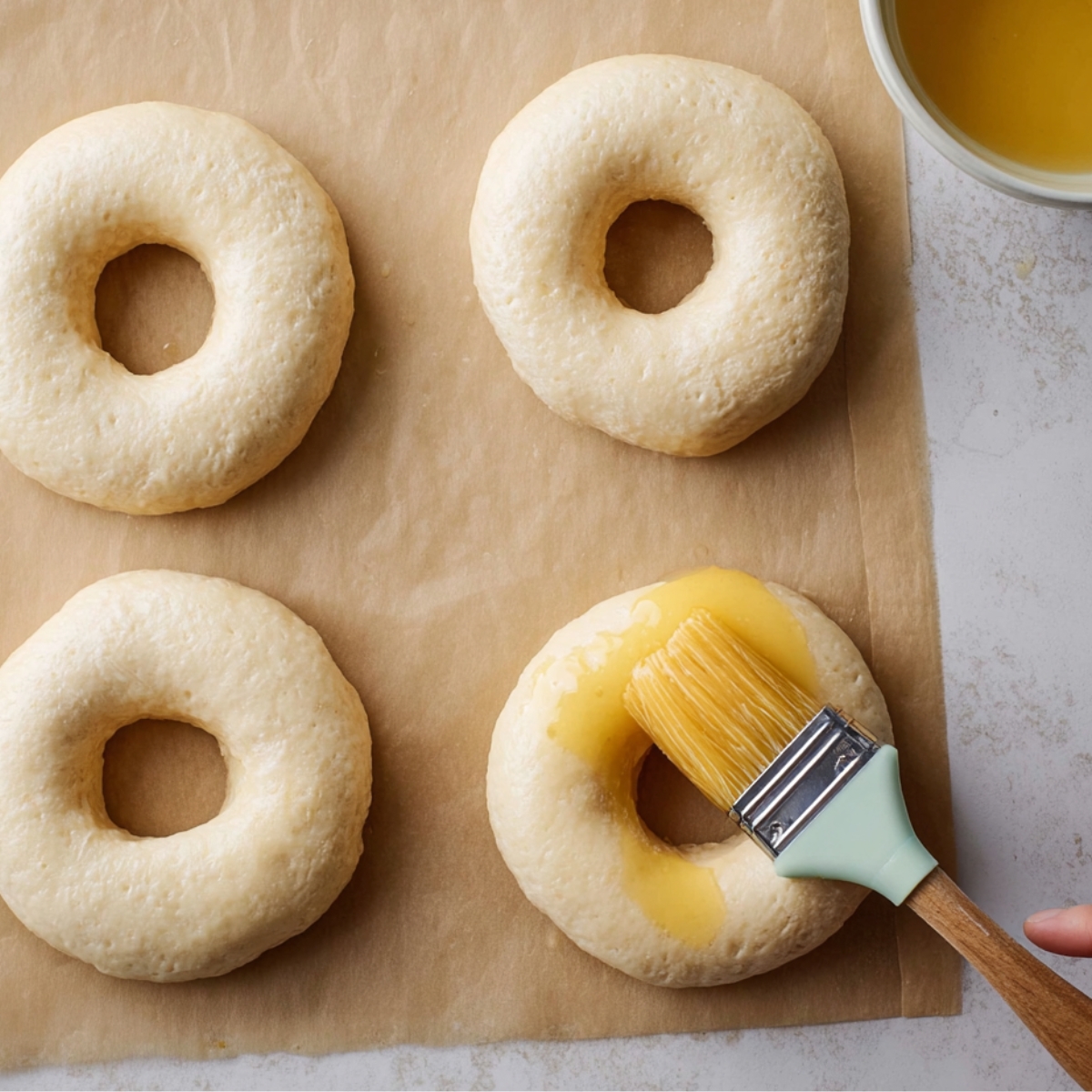 Unbaked bagels on parchment paper being brushed with egg wash, ready for baking, as part of making savory spinach and cottage cheese bagels.