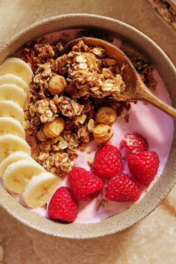 A wholesome homemade high-protein breakfast bowl featuring creamy pink yogurt, crunchy granola, sliced bananas, fresh raspberries, and hazelnuts, perfect for a healthy and energizing start to your day.