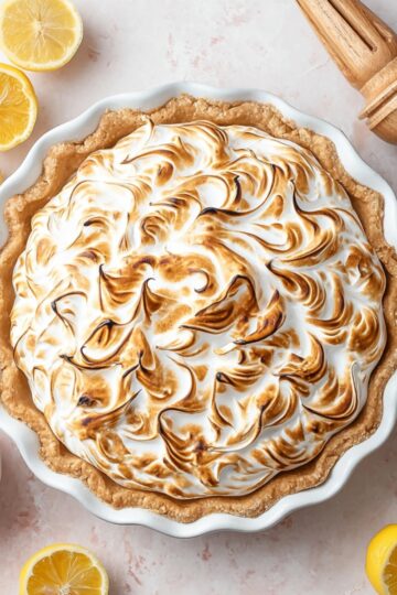 Freshly baked lemon meringue pie with perfectly toasted peaks of meringue, a smooth lemon filling, and a golden flaky crust, surrounded by fresh lemon halves and a small bowl of lemon curd.