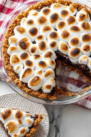 A slice of rich chocolate s’mores brownie pie topped with golden toasted marshmallows in a graham cracker crust, served on a white plate next to the remaining pie. Perfect s’mores dessert recipe.
