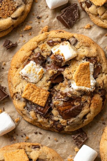 Close-up of freshly baked S’mores cookies on parchment paper, loaded with melted chocolate chunks, toasted marshmallows, and graham cracker pieces, surrounded by scattered chocolate and marshmallows.