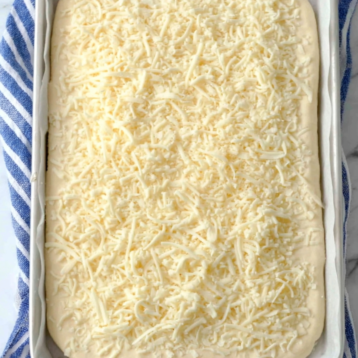 Unbaked pizza dough in a baking tray, generously topped with shredded mozzarella cheese, ready to make homemade Copycat Pizza Hut cheese sticks.