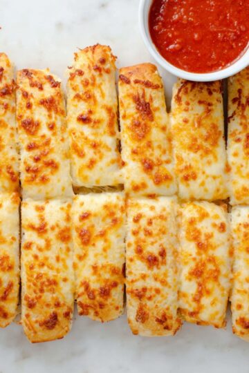 Golden, cheesy breadsticks baked to perfection, served with a small bowl of marinara sauce, homemade Copycat Pizza Hut style cheese sticks recipe.