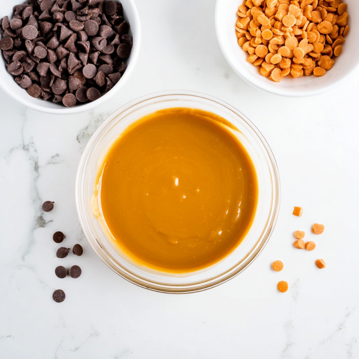 Overhead shot of ingredients for homemade Twix Cookies: a bowl of chocolate chips, a bowl of butterscotch chips, and a bowl of smooth caramel for baking.